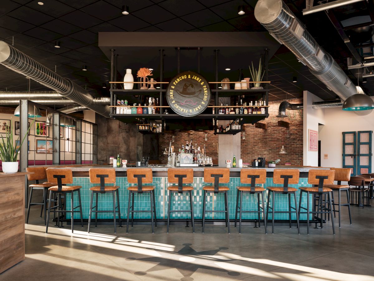 A stylish bar area with a central counter, high chairs, modern decor, and industrial elements like exposed ducts and brick walls.