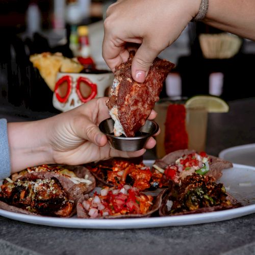 Two hands enjoying a plate of tacos with various fillings and dipping sauces, set on a countertop in a cozy dining setting.