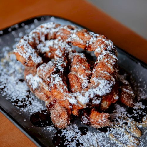 The image shows a plate of churros sprinkled with powdered sugar, served on a rectangular black dish, placed on a wooden surface.