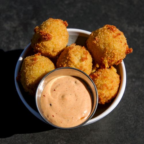 Five golden-brown fried balls are in a white bowl, accompanied by a small cup of creamy dipping sauce in the center.