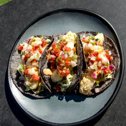 The image shows a plate with three tacos filled with shrimp, diced vegetables, and garnishes on blue tortillas, placed on a dark surface.