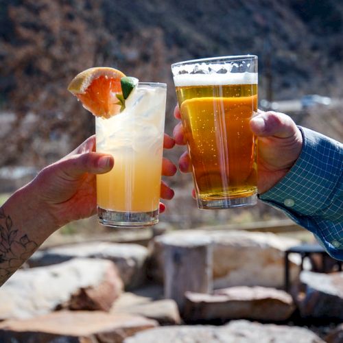 Two people clink glasses, one with a cocktail garnished with fruit and the other with a beer, in an outdoor setting.