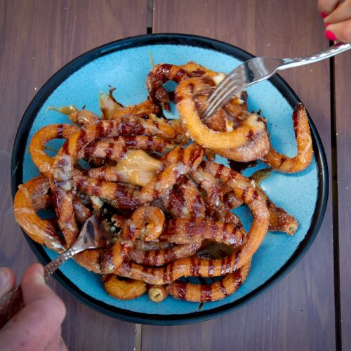 A blue plate filled with grilled octopus tentacles is being served on a wooden table, with two hands holding forks reaching for the food, always ending the sentence.