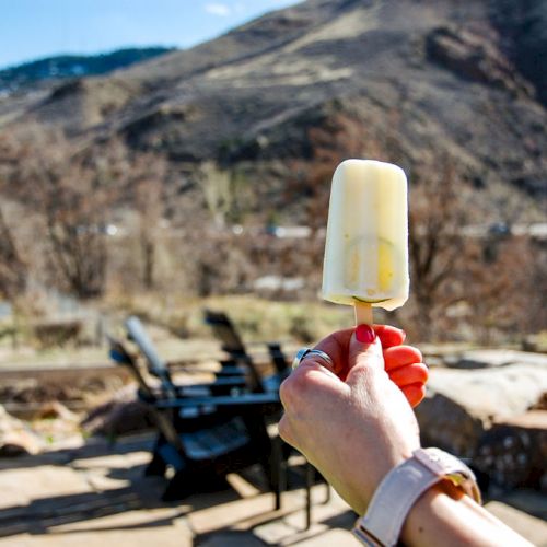 A person holding a popsicle outside, with mountainous scenery and patio furniture in the background.