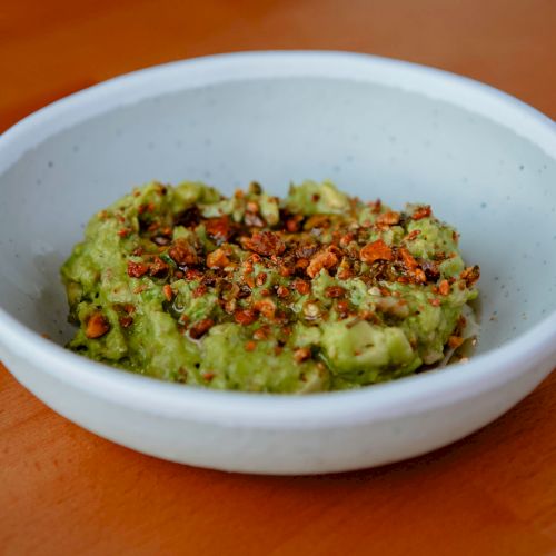 A white ceramic bowl filled with green avocado dip, garnished with seeds and spices, sits on a wooden surface.