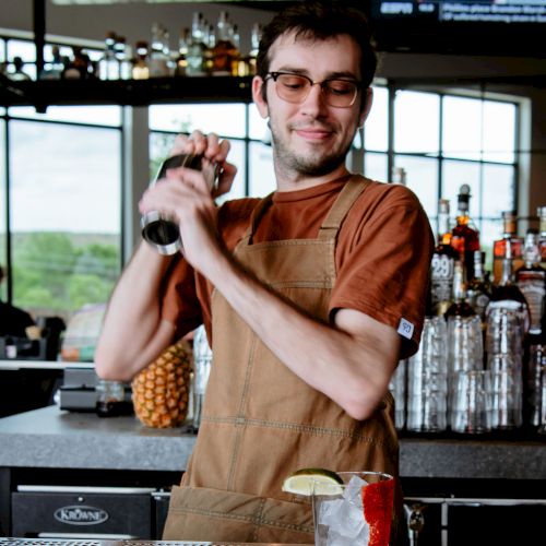 A bartender stands behind the bar, shaking a cocktail shaker with a smile, while a drink with a lime garnish sits on the counter in front of him.