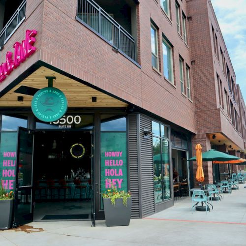 The image shows a corner of a brick building with outdoor seating for a restaurant. The restaurant has colorful signage and potted plants outside.