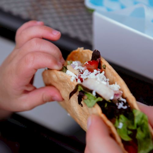 A close-up of a person holding a taco filled with various ingredients like lettuce, cheese, and tomatoes, being handed to or shared with another person.