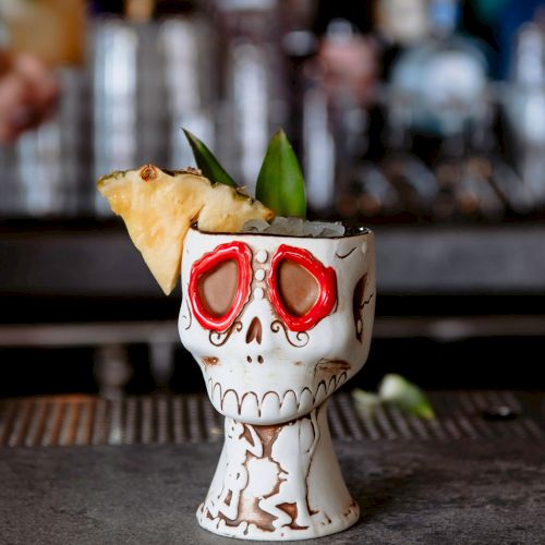 A festive skull-shaped cup with a pineapple slice and leafy garnish sits on a bar counter, with blurry bottles in the background.