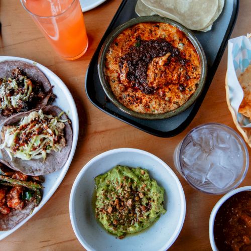 The image shows a spread of Mexican food including tacos, guacamole, salsa, chips, a drink, and a baked dish, all arranged on a wooden table.