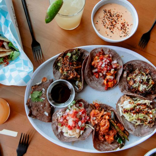 A spread of tacos with various toppings, a bowl of sauce, drink, and utensils on a wooden table, ready to be enjoyed.