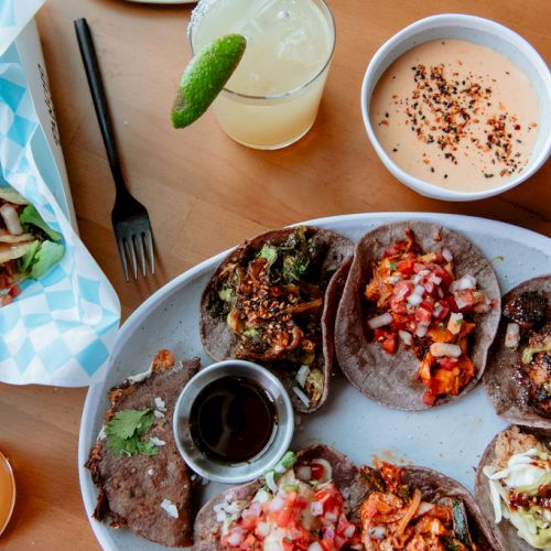 The image shows a plate of assorted tacos, a bowl of dipping sauce, drinks with lime, and a basket of tacos on a wooden table, with forks.