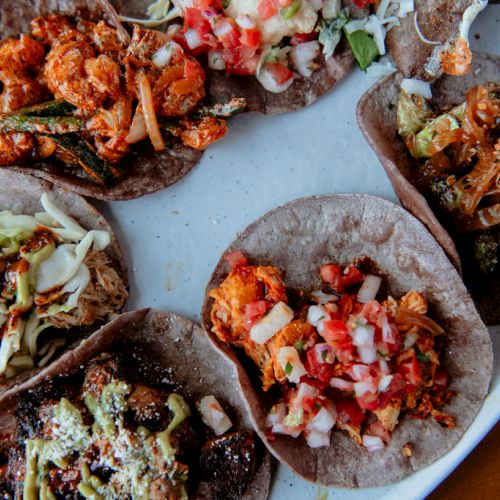 A plate of six tacos with various toppings including diced tomatoes, onions, greens, and sauces.