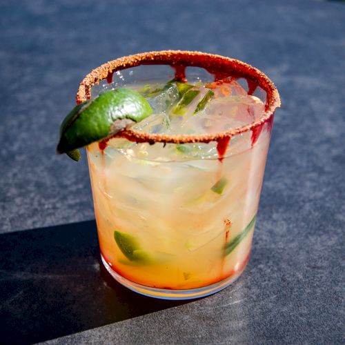 A refreshing cocktail with ice, lime, and a chili-powder-rimmed glass, placed on a dark surface.