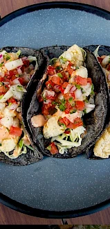 The image shows three tacos with a colorful topping, placed on a dark plate. The toppings appear to be a mix of vegetables and possibly seafood.