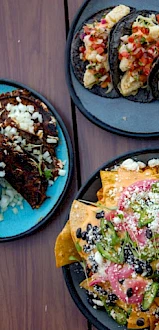 The image shows plates of nachos with toppings, dark-colored tacos filled with various ingredients, and a blue plate with another taco variety.