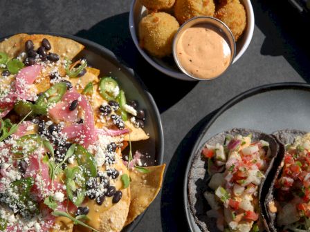 The image shows a plate of nachos with toppings, a bowl of fried croquettes with sauce, and two tacos with mixed fillings on a gray plate.