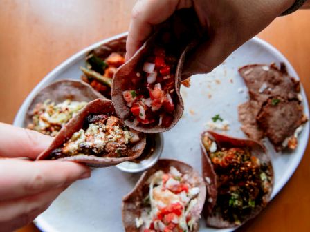 The image shows various tacos on a plate, with two hands holding up tacos filled with ingredients like salsa, onions, and meat.